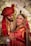 A joyful couple is dressed in traditional wedding attire, featuring intricate patterns and rich colors. The bride is adorned with elaborate jewelry including a maang tikka and earrings. The groom wears a patterned red turban. They are surrounded by a floral backdrop, with expressions of happiness as they look towards each other.