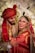 A joyful couple is dressed in traditional wedding attire, featuring intricate patterns and rich colors. The bride is adorned with elaborate jewelry including a maang tikka and earrings. The groom wears a patterned red turban. They are surrounded by a floral backdrop, with expressions of happiness as they look towards each other.