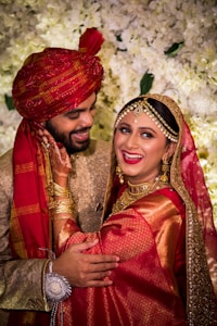 A joyful couple is dressed in traditional wedding attire, featuring intricate patterns and rich colors. The bride is adorned with elaborate jewelry including a maang tikka and earrings. The groom wears a patterned red turban. They are surrounded by a floral backdrop, with expressions of happiness as they look towards each other.