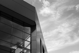 A modern building facade with large, reflective glass windows positioned against a partially cloudy sky. The architecture features clean lines and a sharp, angular design.