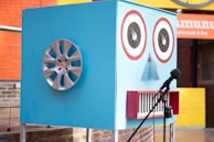 A large blue box resembling a robot face is designed with two circular speaker eyes, a small triangular mesh for a nose, and a rectangular mouth with red border lines. A car wheel hubcap is attached to one side of the box, possibly representing an ear. There is a microphone with a stand positioned in front of the box. The background features a colorful outdoor setting with orange and yellow walls and a restaurant sign.