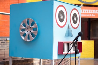 A large blue box resembling a robot face is designed with two circular speaker eyes, a small triangular mesh for a nose, and a rectangular mouth with red border lines. A car wheel hubcap is attached to one side of the box, possibly representing an ear. There is a microphone with a stand positioned in front of the box. The background features a colorful outdoor setting with orange and yellow walls and a restaurant sign.