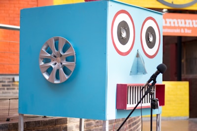 A large blue box resembling a robot face is designed with two circular speaker eyes, a small triangular mesh for a nose, and a rectangular mouth with red border lines. A car wheel hubcap is attached to one side of the box, possibly representing an ear. There is a microphone with a stand positioned in front of the box. The background features a colorful outdoor setting with orange and yellow walls and a restaurant sign.