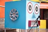 A large blue box resembling a robot face is designed with two circular speaker eyes, a small triangular mesh for a nose, and a rectangular mouth with red border lines. A car wheel hubcap is attached to one side of the box, possibly representing an ear. There is a microphone with a stand positioned in front of the box. The background features a colorful outdoor setting with orange and yellow walls and a restaurant sign.
