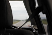 A view from inside a car looking out the front windshield. The interior features a seat with a headrest and a visible seatbelt in use. The road outside is partially visible, slightly blurred, suggesting motion or focus on the interior elements.