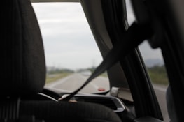 A view from inside a car looking out the front windshield. The interior features a seat with a headrest and a visible seatbelt in use. The road outside is partially visible, slightly blurred, suggesting motion or focus on the interior elements.