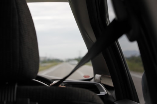 A view from inside a car looking out the front windshield. The interior features a seat with a headrest and a visible seatbelt in use. The road outside is partially visible, slightly blurred, suggesting motion or focus on the interior elements.