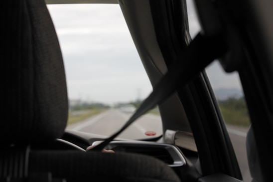 A view from inside a car looking out the front windshield. The interior features a seat with a headrest and a visible seatbelt in use. The road outside is partially visible, slightly blurred, suggesting motion or focus on the interior elements.