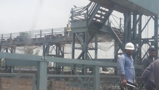 A group of workers wearing helmets stands near a metal bridge structure. The bridge features multiple beams and a staircase, with netting and wires visible. One worker holds a tool, possibly a construction instrument.