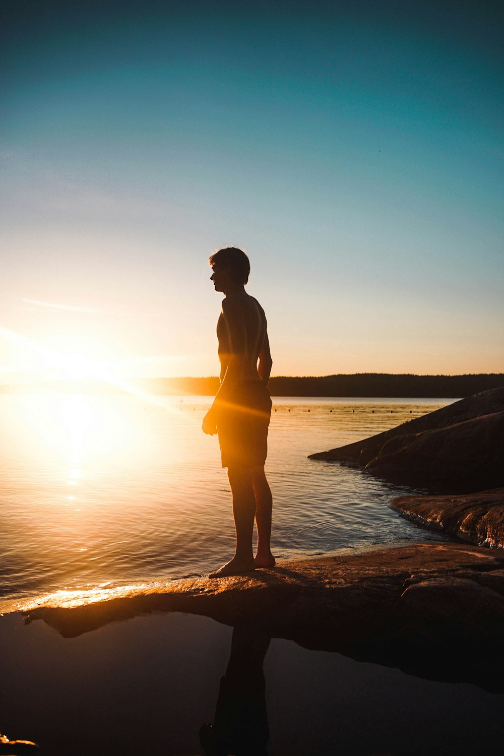 man standing on rock besides body of water during sunset