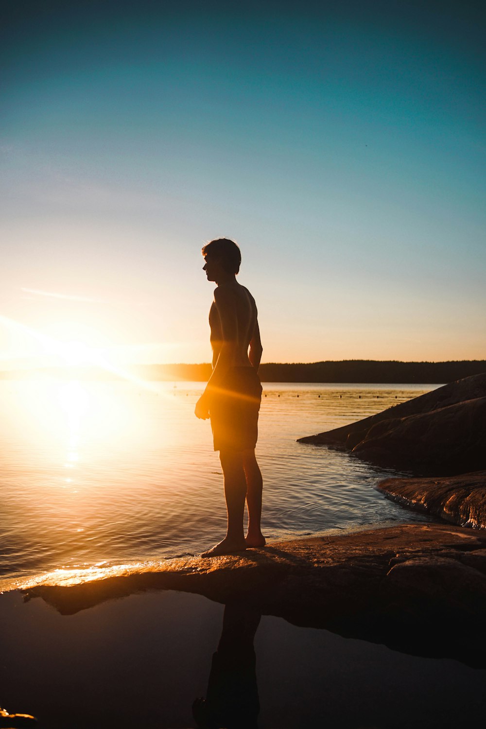 uomo in piedi sulla roccia oltre allo specchio d'acqua durante il tramonto