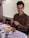 A person is sitting at a table, smiling while holding chopsticks. In front of them is a plate filled with a variety of foods, including sliced cucumbers, cabbage, tomatoes, mushrooms, and beans. A cup of tea and a spoon are on the table, which is covered with a checkered tablecloth. The background features blinds partially covering a window.