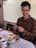 A person is sitting at a table, smiling while holding chopsticks. In front of them is a plate filled with a variety of foods, including sliced cucumbers, cabbage, tomatoes, mushrooms, and beans. A cup of tea and a spoon are on the table, which is covered with a checkered tablecloth. The background features blinds partially covering a window.