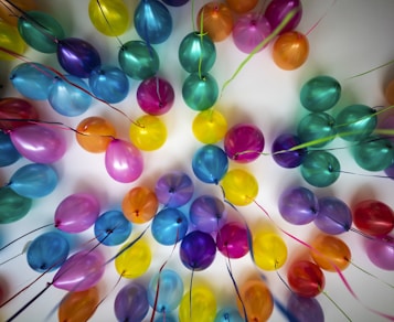 A vibrant array of colorful balloons is attached to the ceiling. These balloons come in shades of blue, purple, yellow, green, and pink, creating a lively and festive atmosphere. Each balloon is tied with a string, contributing to the overall aesthetic.