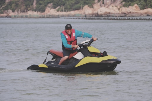 A person wearing a red life vest and a blue long-sleeved shirt is riding a yellow and black jet ski on a body of water. They appear to be enjoying the activity, and their expression suggests joy or excitement. The background features a shoreline with trees and rocks.