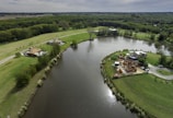 An aerial view of a serene landscape with a river or lake surrounded by lush green fields and dense forests. The image includes a couple of houses, one under construction, situated on the banks. A road passes by the left, edged with trees and grass patches. The water reflects the cloudy sky above.