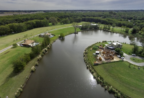An aerial view of a serene landscape with a river or lake surrounded by lush green fields and dense forests. The image includes a couple of houses, one under construction, situated on the banks. A road passes by the left, edged with trees and grass patches. The water reflects the cloudy sky above.