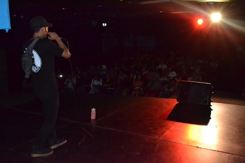 A person is performing on stage with a microphone, facing a seated audience in a dimly lit space. The lighting is focused on the performer, creating a contrast with the darker background. An orange-red spotlight illuminates part of the stage.