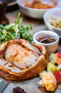 A dish featuring a rolled roast with a crispy outer layer and marbled interior is surrounded by various accompaniments. There is a side of fresh green salad and a small white bowl containing a dark sauce with a spoon. Additionally, some assorted pickled vegetables, including cauliflower and peppers, add colorful elements to the composition.
