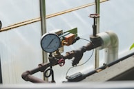 A complex network of industrial pipes and gauges is arranged against a translucent wall. The dominant feature is a pressure gauge reading between 20 and 30, connected to black and rust-colored metal pipes. A red valve and an orange electrical component are attached to the structure, adding to the industrial feel.