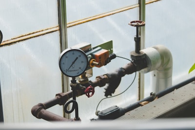 A complex network of industrial pipes and gauges is arranged against a translucent wall. The dominant feature is a pressure gauge reading between 20 and 30, connected to black and rust-colored metal pipes. A red valve and an orange electrical component are attached to the structure, adding to the industrial feel.