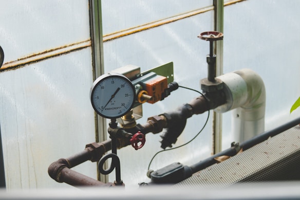A complex network of industrial pipes and gauges is arranged against a translucent wall. The dominant feature is a pressure gauge reading between 20 and 30, connected to black and rust-colored metal pipes. A red valve and an orange electrical component are attached to the structure, adding to the industrial feel.