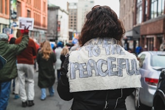 A person with long curly hair is wearing a large fabric sign that reads 'POWER TO THE PEACEFUL' on their back. They are part of a crowd walking down a city street. People around are holding signs, indicating a peaceful protest or march. Buildings line the street on either side, and parked cars are visible.