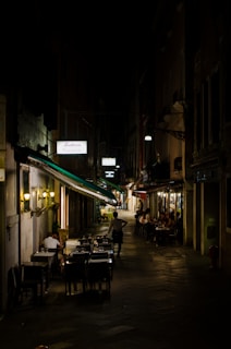 A narrow, dimly lit street with outdoor seating at two restaurants, occupied by people dining and socializing under warm lighting. The street appears to be in a European city, with a quaint and intimate atmosphere enhanced by the dark surroundings and cozy setting.