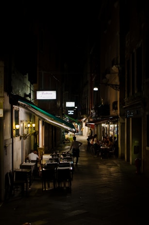 A narrow, dimly lit street with outdoor seating at two restaurants, occupied by people dining and socializing under warm lighting. The street appears to be in a European city, with a quaint and intimate atmosphere enhanced by the dark surroundings and cozy setting.
