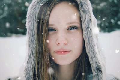 A close-up of a person wearing a hooded fur coat with a background of falling snowflakes. The individual has long straight hair and blue eyes, and the expression appears calm and contemplative. The background is blurred, emphasizing the falling snow and wintry atmosphere.