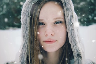 A close-up of a person wearing a hooded fur coat with a background of falling snowflakes. The individual has long straight hair and blue eyes, and the expression appears calm and contemplative. The background is blurred, emphasizing the falling snow and wintry atmosphere.