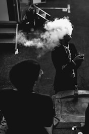 A person in a suit is exhaling a large plume of smoke that obscures their face. The setting appears to be outdoors with a table in the foreground holding a bottle. Another person is partially visible in silhouette.