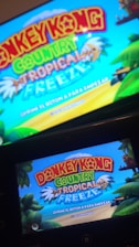 A television screen and a handheld device display the title screen of a video game, featuring vibrant text 'Donkey Kong Country Tropical Freeze' against a bright blue sky. The background includes tropical elements such as palm trees and lush greenery.