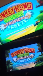 A television screen and a handheld device display the title screen of a video game, featuring vibrant text 'Donkey Kong Country Tropical Freeze' against a bright blue sky. The background includes tropical elements such as palm trees and lush greenery.