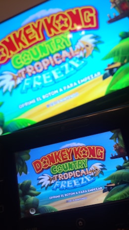 A television screen and a handheld device display the title screen of a video game, featuring vibrant text 'Donkey Kong Country Tropical Freeze' against a bright blue sky. The background includes tropical elements such as palm trees and lush greenery.