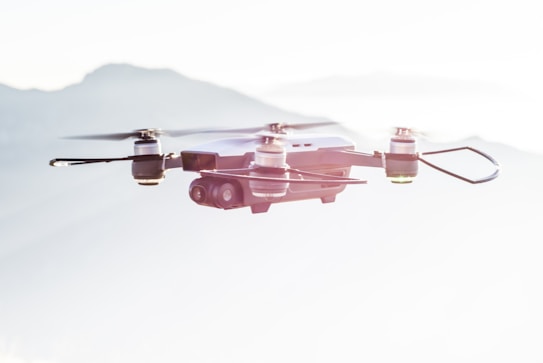 A quadcopter drone flying against a backdrop of soft, blurred mountains. The drone has four propellers and a camera mounted on its underside.