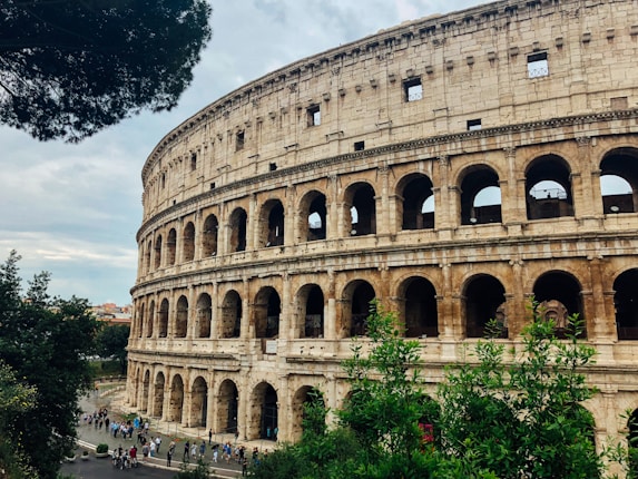 The ancient Colosseum stands majestically against a partly cloudy sky, surrounded by greenery and a bustling crowd of tourists. Its iconic arches and weathered stone facade are clearly visible, capturing the grandeur of Roman architecture.