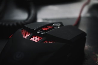 A close-up view of a sleek, black gaming mouse with red accents and buttons. The mouse design includes a textured grip and a scroll wheel, with a coiled wire partially visible in the background.