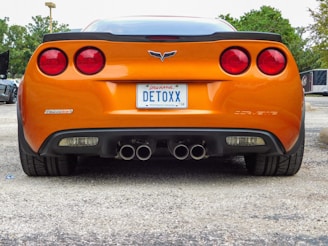 An orange sports car is parked with a rear view showing its sleek design. The car has dual exhaust pipes and a personalized license plate that reads 'DETOXX'. The background includes trees and other parked vehicles.