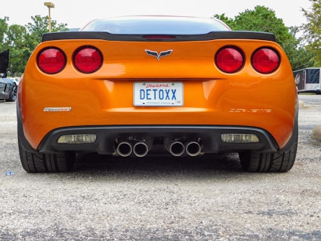 An orange sports car is parked with a rear view showing its sleek design. The car has dual exhaust pipes and a personalized license plate that reads 'DETOXX'. The background includes trees and other parked vehicles.