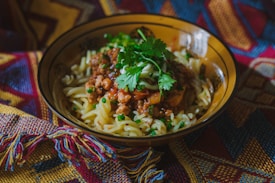 A bowl of pasta topped with a savory meat sauce and garnished with fresh cilantro leaves. The background features a colorful, patterned fabric with geometric designs, adding a warm and homely feel to the setting.