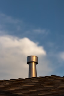 A metallic chimney vent extends from the peak of a shingled roof against a backdrop of a partly cloudy sky. The texture of the roof shingles is visible, and the chimney vent reflects light, giving it a metallic sheen.