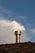 A metallic chimney vent extends from the peak of a shingled roof against a backdrop of a partly cloudy sky. The texture of the roof shingles is visible, and the chimney vent reflects light, giving it a metallic sheen.