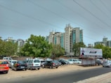 A cityscape with modern apartment buildings in the background, a variety of parked cars in a lot, and a large billboard advertising design institute courses. Green trees are scattered among the parked vehicles, and the sky is overcast with visible power lines.