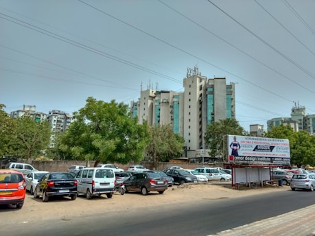 A cityscape with modern apartment buildings in the background, a variety of parked cars in a lot, and a large billboard advertising design institute courses. Green trees are scattered among the parked vehicles, and the sky is overcast with visible power lines.