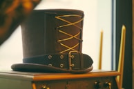 A vintage leather top hat with a decorative lace-up design rests on a wooden cabinet. The hat features metal eyelets and a buckle strap, adding to its antique charm. In the background, there are two tall candles, out of focus, near a window, creating a warm ambiance.