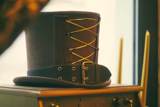 A vintage leather top hat with a decorative lace-up design rests on a wooden cabinet. The hat features metal eyelets and a buckle strap, adding to its antique charm. In the background, there are two tall candles, out of focus, near a window, creating a warm ambiance.
