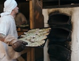 A person wearing a white bandana and shirt is holding a large tray of flatbreads covered with herbs and preparing to place them into a rustic, stone oven.