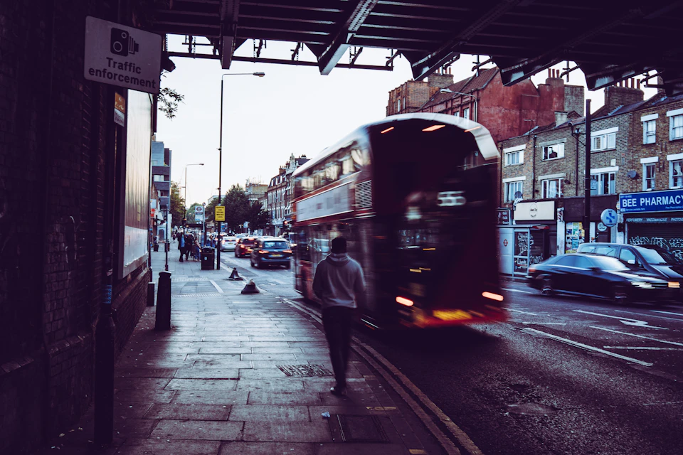 The image depicts a busy street scene with a red double-decker bus driving down the road, surrounded by various vehicles such as cars and trucks. A pedestrian is seen walking on the sidewalk next to the bus, adding life to the urban setting. The perspective of the photo is taken from under an overpass, providing a unique viewpoint that captures both the bustling activity and the architectural elements of the cityscape.

There are several people visible in the image, some walking along the sidewalk or waiting at the bus stop, contributing to the dynamic atmosphere of the scene. A traffic light can be seen further down the street, indicating the presence of an intersection where vehicles and pedestrians share the space.