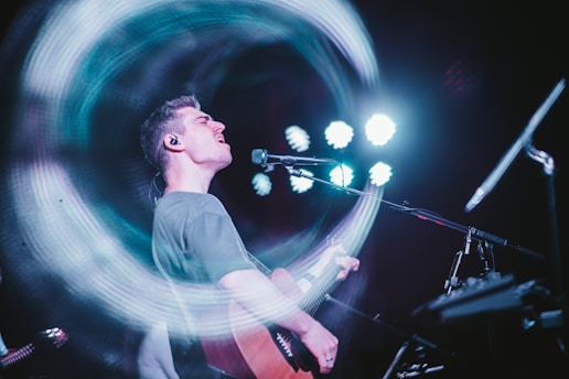 A musician passionately singing and playing an acoustic guitar on stage. There's a dynamic motion effect with swirling lights around the performer, while bright spotlights shine in the background.